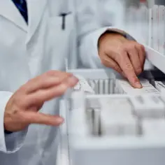 Farmaceuta szuka zamiennika leku. Dłonie w białym fartuchu przeglądają opakowania leków w aptece.