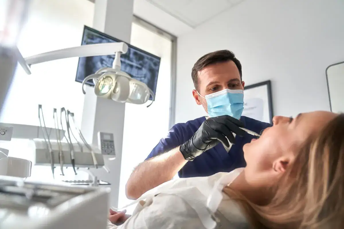 Dentysta w masce wykonuje szynowanie zębów pacjentce. W tle widać aparat RTG i lampę zabiegową.